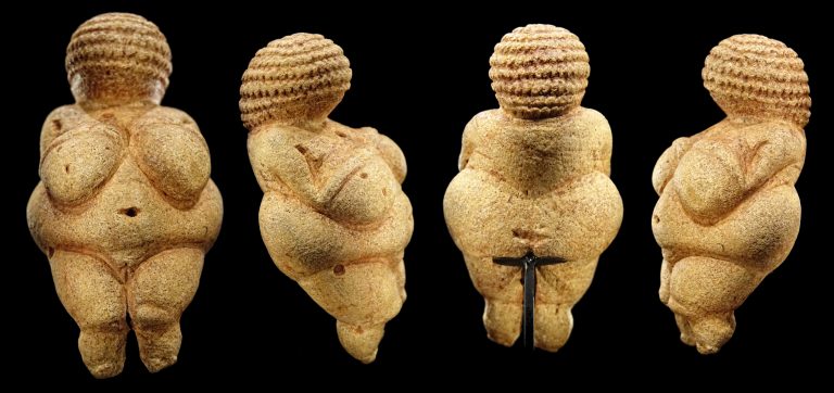A female Venus figure with exaggerated sexual characteristics. The head is relatively small and lacks facial features, while the body is depicted with ample breasts, a round belly, and wide hips.