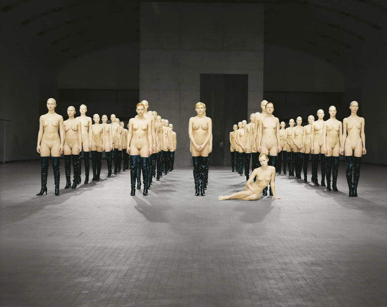 staged installation with multiple nude female models standing or seated motionless in a gallery space, arranged in a formal, sculptural composition