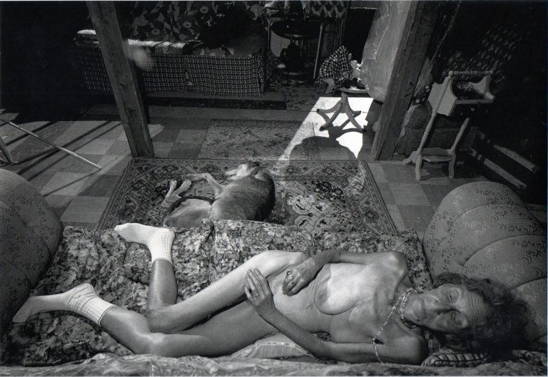 Photographic portrait of an elderly nude woman with exposed breasts and pubic area, laying in a stark, intimate interior with unidealized, raw realism.