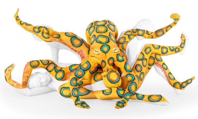 Body-painting photograph of a nude model whose body is painted and posed so limbs merge into tentacle-like forms, creating the illusion of an octopus in a studio setting.
