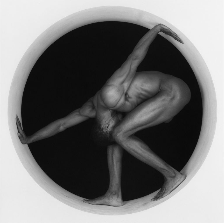 black-and-white studio portrait of a nude male figure standing in a composed, sculptural pose, with controlled lighting emphasizing body form and musculature