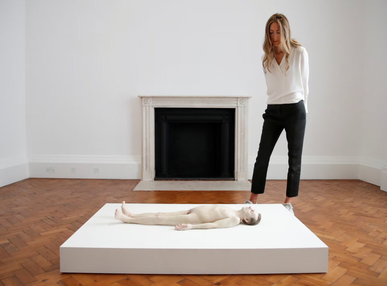 Hyperrealistic sculpture of a naked male body lying on the ground, with exposed genitals, shrunken scale, and detailed skin textures emphasizing lifelessness and vulnerability.