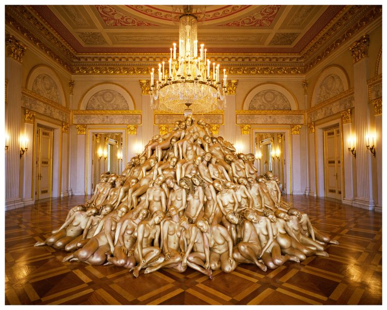 large-scale installation photograph of hundreds of nude participants arranged closely together inside an opera house, forming a dense, coordinated human composition